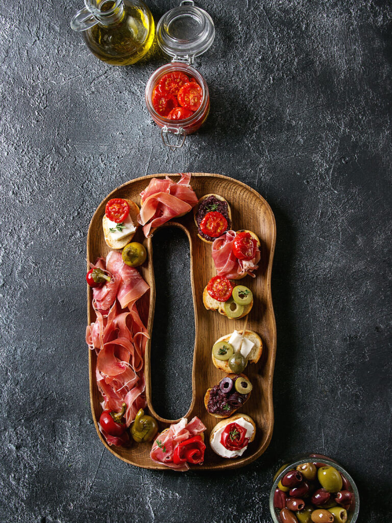 Tapas-Brett mit kleinen Bruschetta, Serrano-Schinken, Oliven und Ofentomaten auf Holzbrett