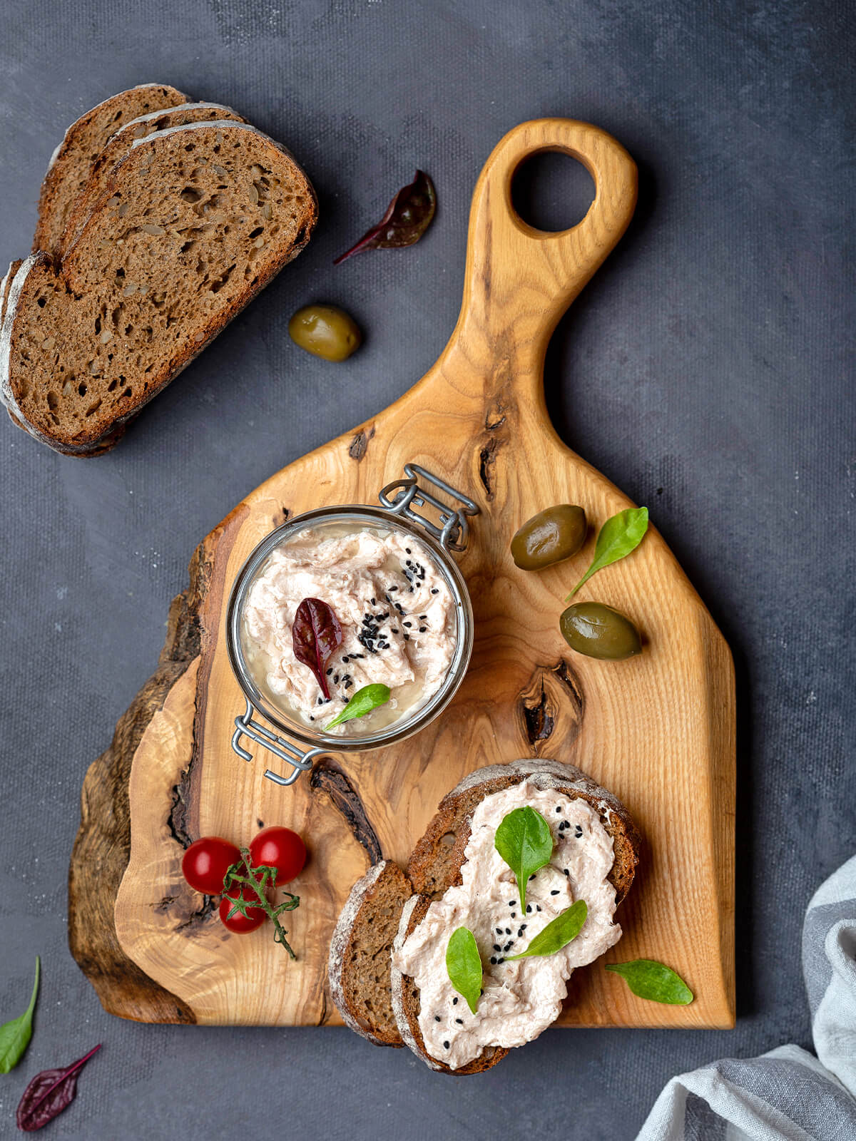 Thunfisch-Rillettes im Bügelglas auf einem Holzbrett mit dunklem Landbrot, grünen Oliven und Cherrytomaten