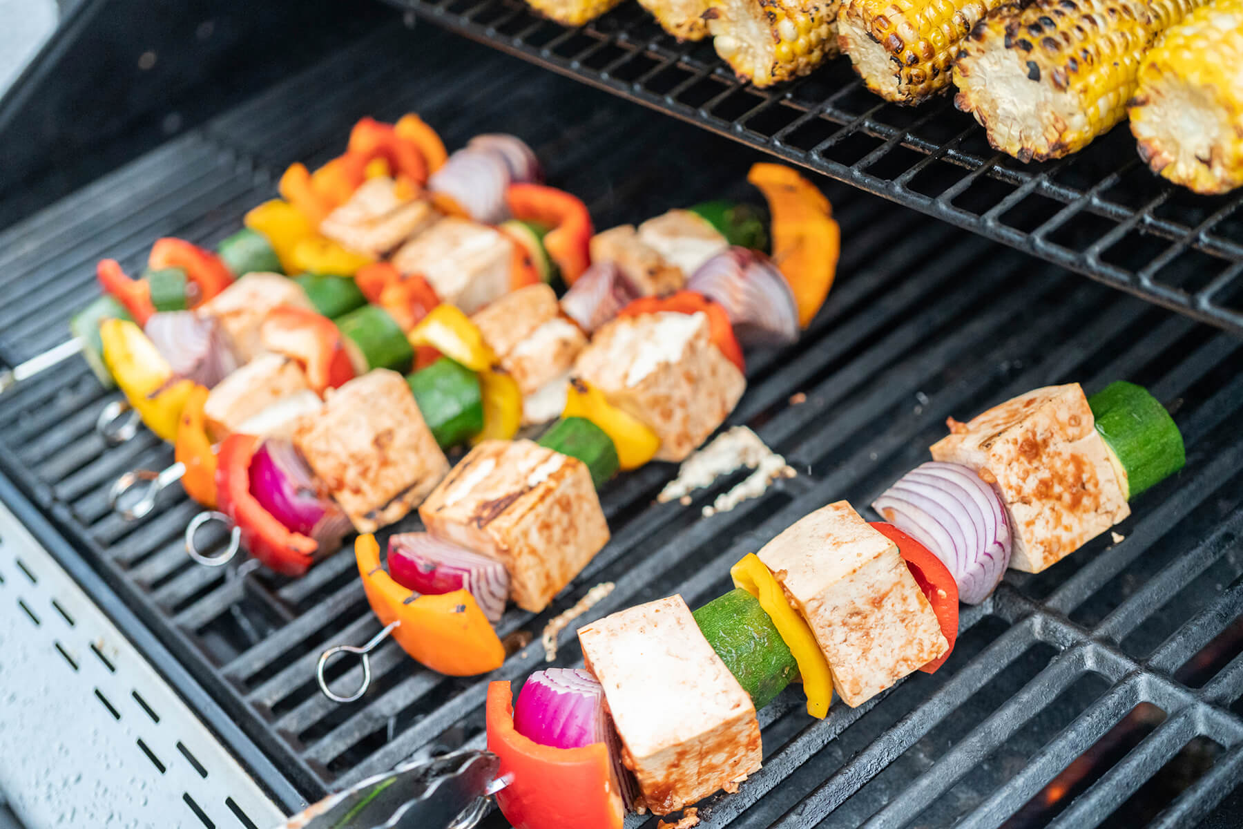 Marinierte Tofu-Spieße mit Paprika und Zucchini auf dem Grillrost, Maiskolben im Hintergrund