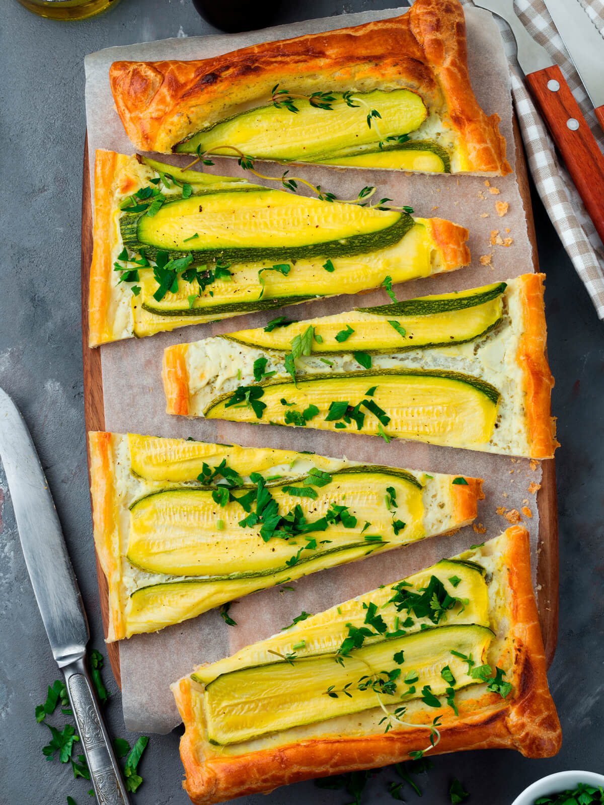 In Stücke geschnittene Zucchini-Ricotta-Tarte aus Blätterteig auf Backpapier mit Messer