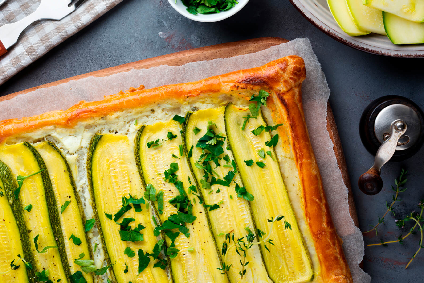 Zucchini-Ricotta-Tarte mit grünen und gelben Zucchini und frischen Kräutern, daneben rohe Zucchini