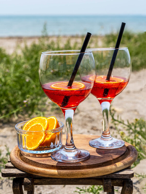 Zwei Gläser alkoholfreier Bitter-Orangen-Aperitif in leuchtendem Rot mit Orangenscheibe und Strohhalm auf einem Holzbrett am Meer