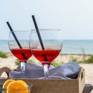 Zwei Gläser alkoholfreier Bitter-Orangen-Aperitif in leuchtendem Rot mit Strohhalmen in einem Holztablett am Strand