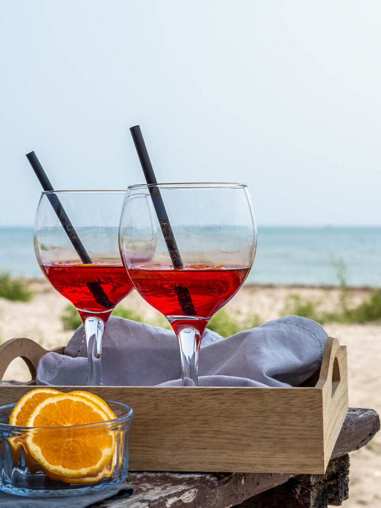 Zwei Gläser alkoholfreier Bitter-Orangen-Aperitif in leuchtendem Rot mit Strohhalmen in einem Holztablett am Strand