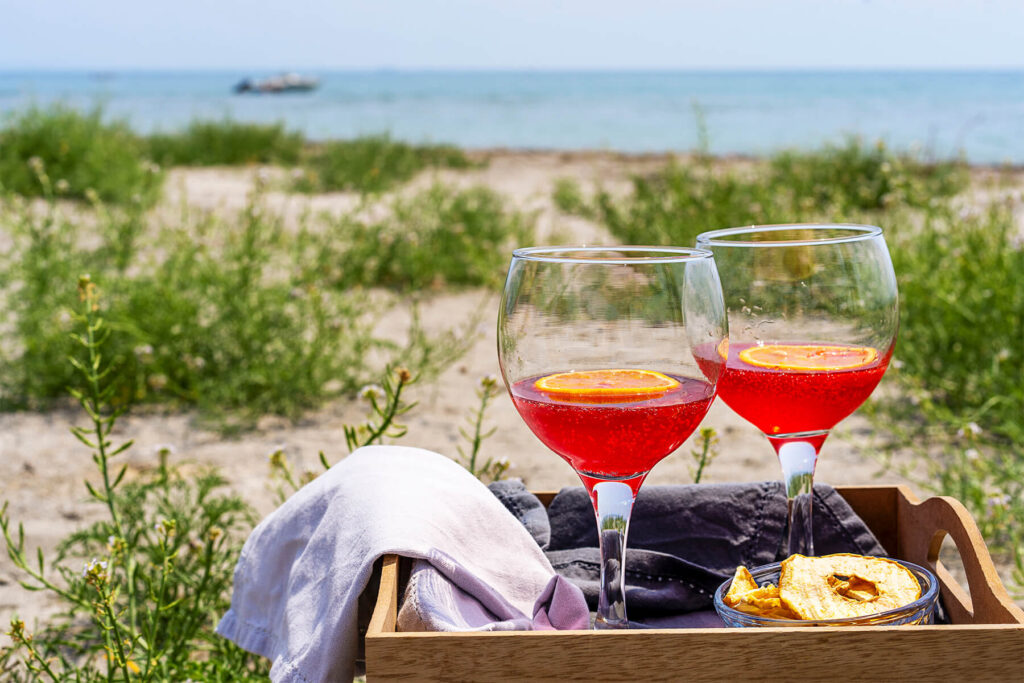 Zwei Gläser alkoholfreier Bitter-Orangen-Aperitif in einem Holztablett mit Tuch in den Dünen, im Hintergrund Meer und ein kleines Boot am Horizont
