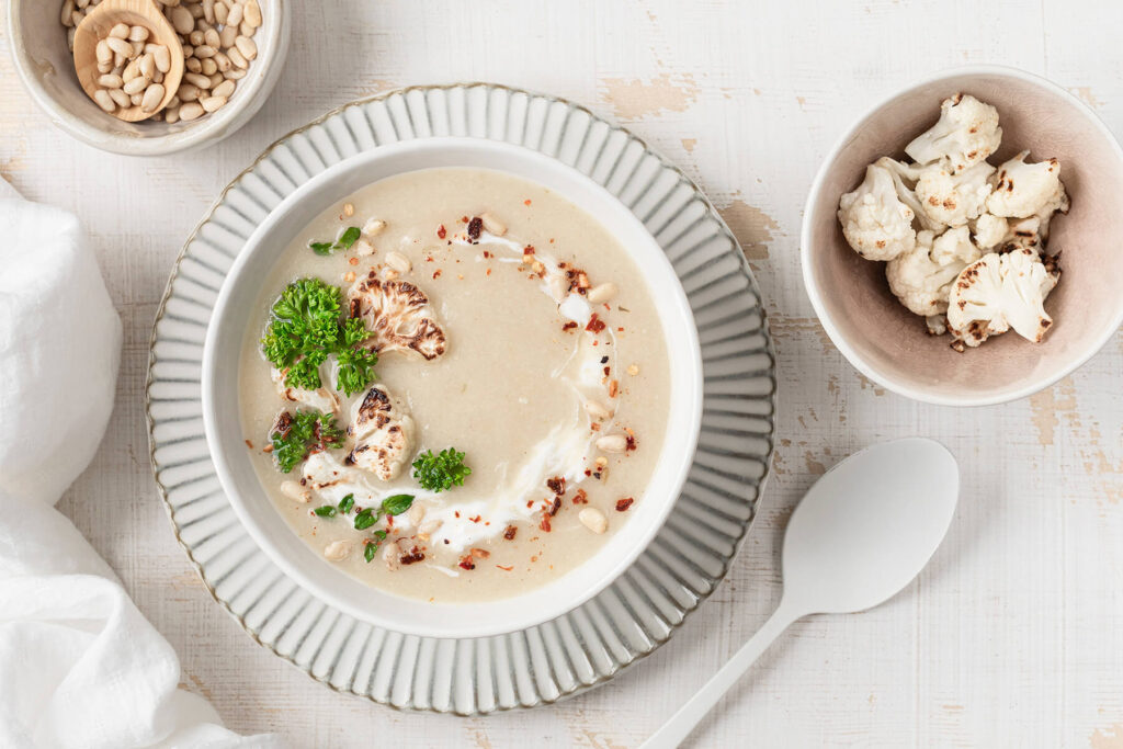 Helle Blumenkohl-Cremesuppe in einer Schale mit Sahne-Strudel, gerösteten Blumenkohl-Röschen, Pinienkernen, frischer Petersilie und ein paar roten Chiliflocken