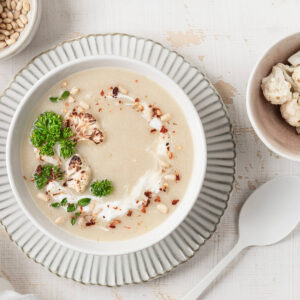 Helle Blumenkohl-Cremesuppe in einer Schale mit Sahne-Strudel, gerösteten Blumenkohl-Röschen, Pinienkernen, frischer Petersilie und ein paar roten Chiliflocken