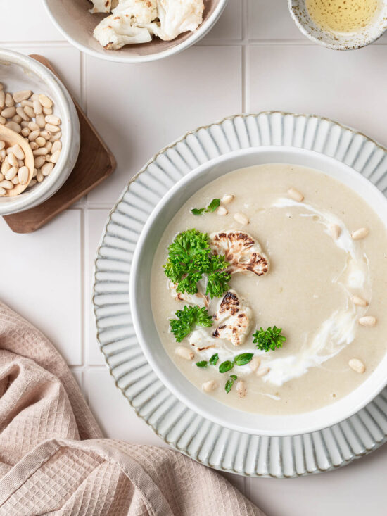 Helle, samtige Blumenkohl-Cremesuppe in einer Schale mit Sahne-Strudel, gerösteten Blumenkohl-Röschen, Pinienkernen und frischer krauser Petersilie