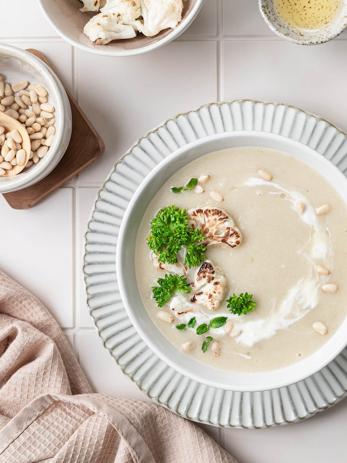 Helle, samtige Blumenkohl-Cremesuppe in einer Schale mit Sahne-Strudel, gerösteten Blumenkohl-Röschen, Pinienkernen und frischer krauser Petersilie