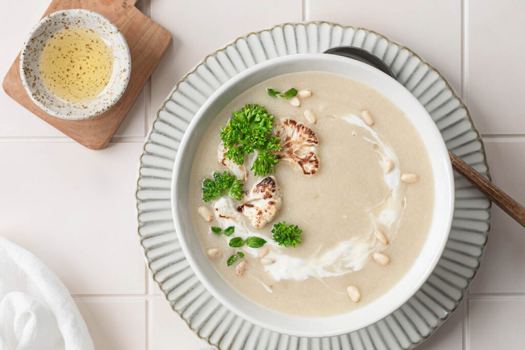 Helle Blumenkohl-Cremesuppe von oben mit Sahne-Strudel, gerösteten Röschen, Pinienkernen und Petersilie, daneben ein Schälchen Öl
