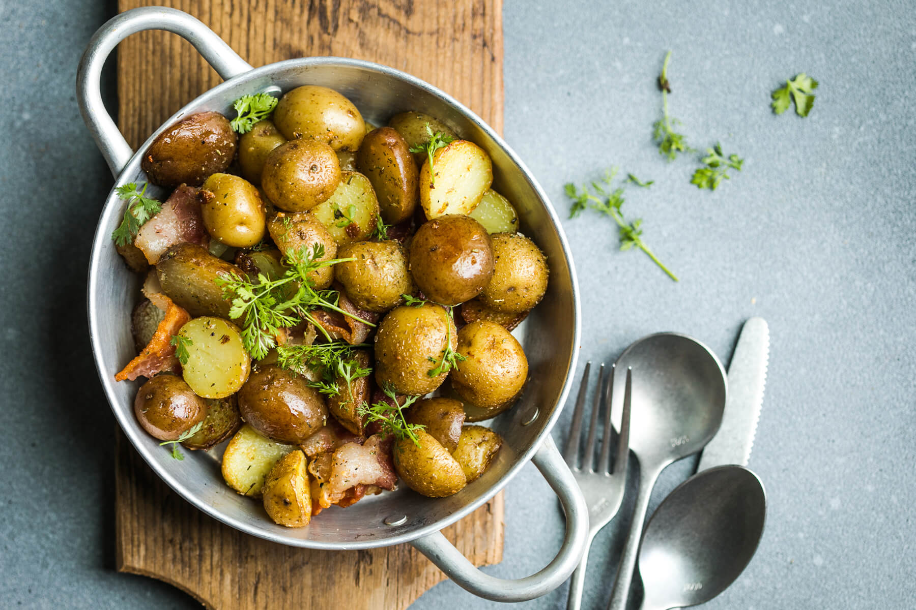 Junge Bratkartoffeln mit Speck und Petersilie in einer Pfanne mit zwei Henkeln von oben, goldbraune kleine Kartoffeln mit Schale und frische Kräuter