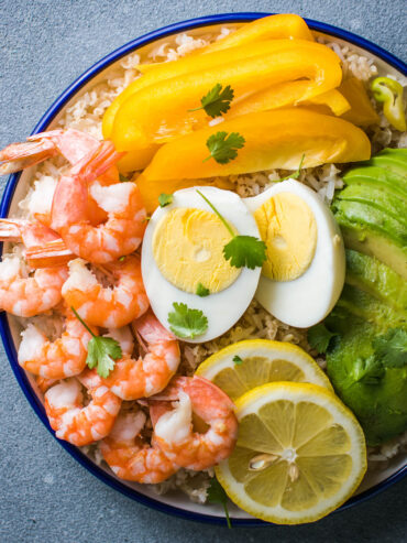 Buddha Bowl mit Avocado, Garnelen und Reis – rosa Garnelen, Avocadofächer, gelbe Paprika, halbiertes Ei und Zitrone auf Vollkornreis in einer blau geränderten Schale