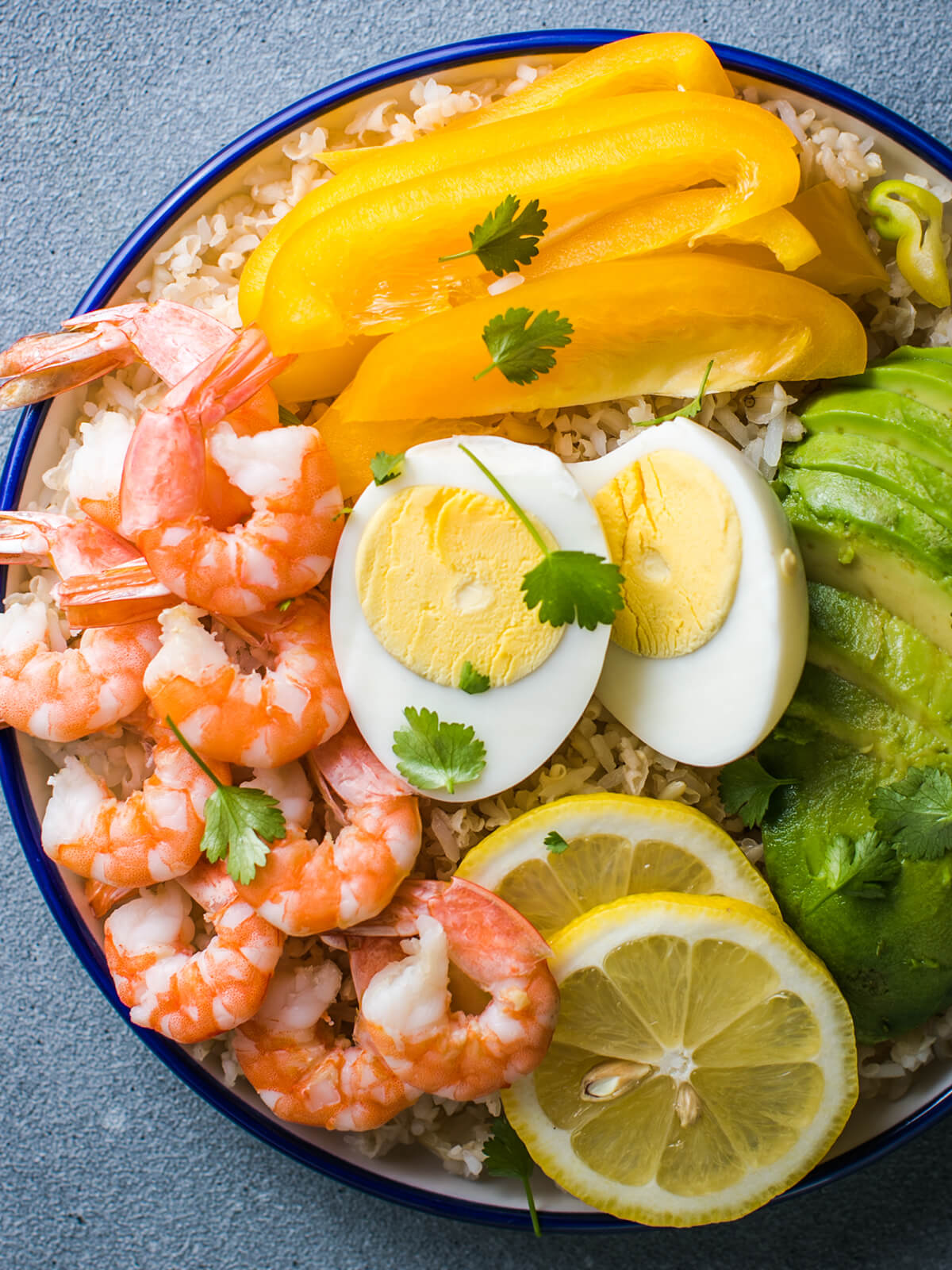 Buddha Bowl mit Avocado, Garnelen und Reis – rosa Garnelen, Avocadofächer, gelbe Paprika, halbiertes Ei und Zitrone auf Vollkornreis in einer blau geränderten Schale