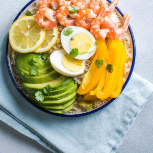 Buddha Bowl mit Avocado, Garnelen und Reis – Garnelen, Avocadofächer, gelbe Paprika, halbiertes Ei und Zitrone auf Vollkornreis in einer blau geränderten Schale