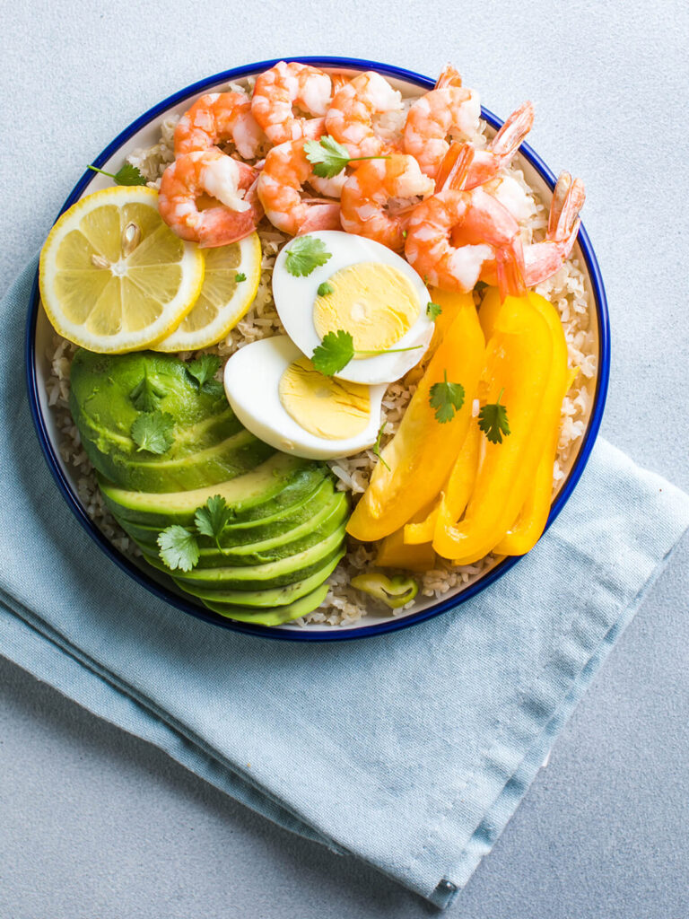 Buddha Bowl mit Avocado, Garnelen und Reis – Garnelen, Avocadofächer, gelbe Paprika, halbiertes Ei und Zitrone auf Vollkornreis in einer blau geränderten Schale