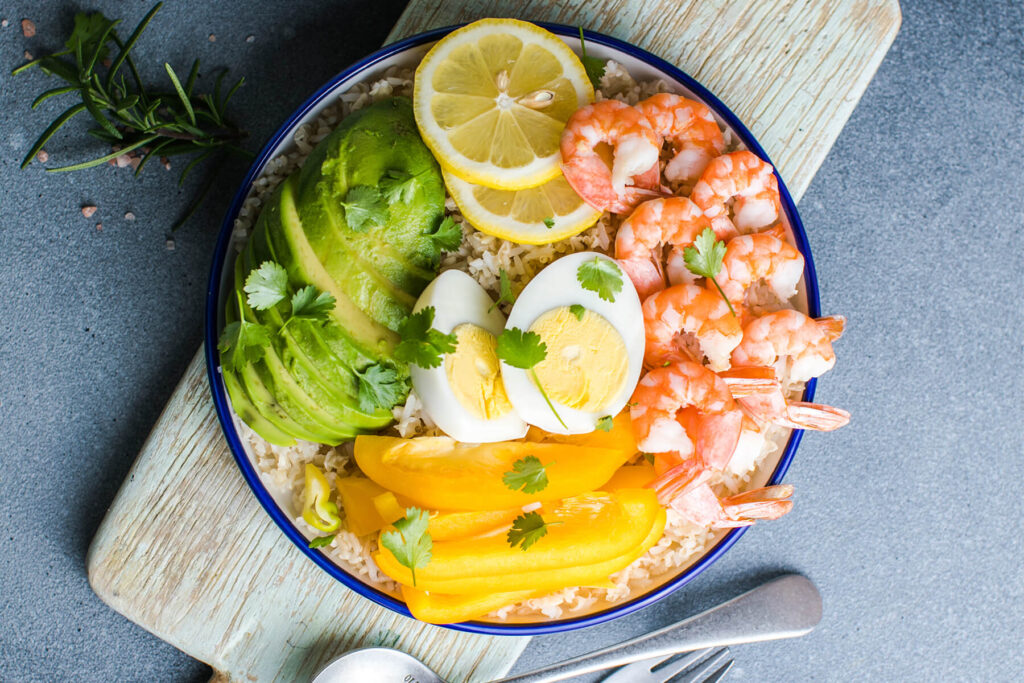 Fertige Buddha Bowl mit Garnelen, Avocadofächern, gelber Paprika und halbiertem Ei auf einem Holzbrett, daneben Besteck