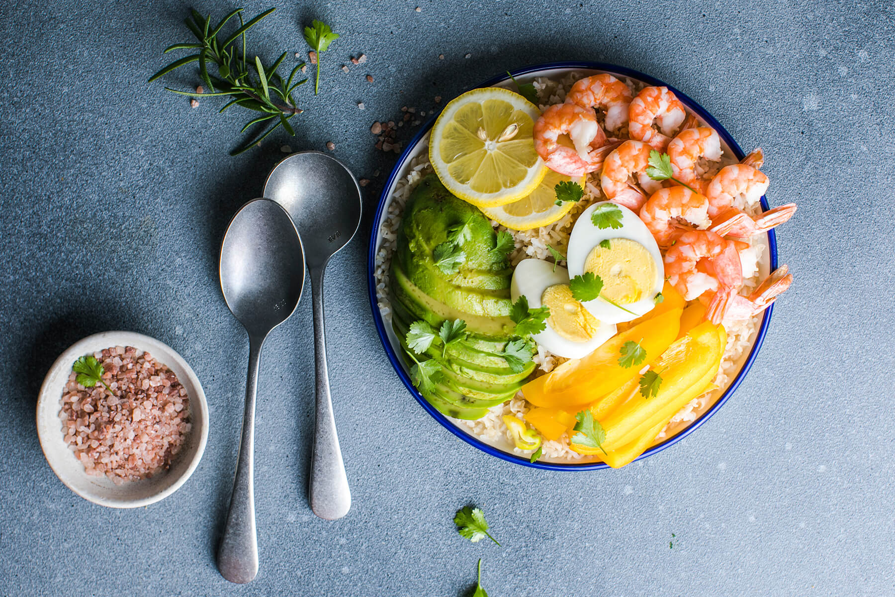 Buddha Bowl mit Garnelen, Avocado, gelber Paprika, halbiertem Ei und Zitrone in einer blau geränderten Schale auf grauem Stein, daneben zwei Löffel, ein Schälchen mit grobem Salz und ein Zweig Rosmarin