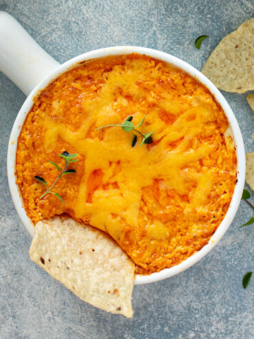 Überbackener Buffalo-Chicken-Dip in einer weißen Schale mit goldenem Cheddar, einem eingetauchten Tortilla-Chip und frischem Oregano