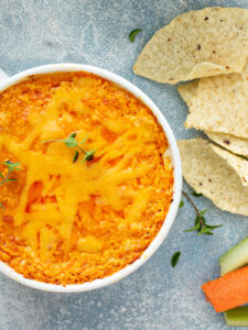 Überbackener Buffalo-Chicken-Dip in einer weißen Schale mit geschmolzenem Cheddar, dazu Tortilla-Chips und Karotten- und Selleriesticks