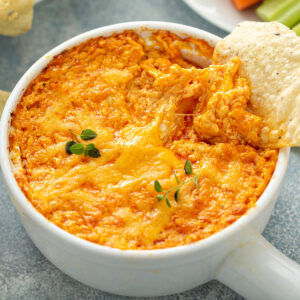 Überbackener Buffalo-Chicken-Dip in einer weißen Schale mit goldenem Cheddar, einem eingetauchten Tortilla-Chip, Karotten- und Selleriesticks