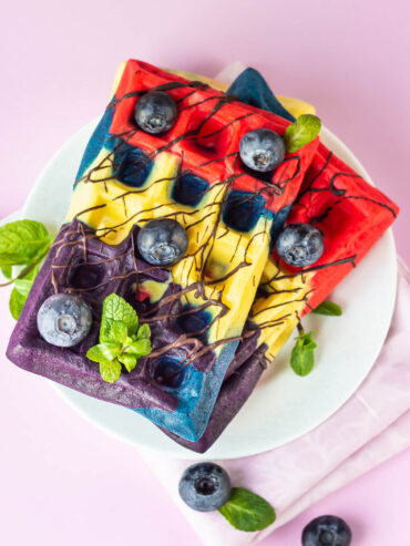 Bunte belgische Waffeln in Rot, Gelb, Blau und Violett, überzogen mit dunklem Schokoladen-Drizzle, frischen Heidelbeeren und Minze auf einem weißen Teller