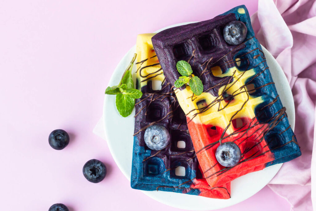 Zwei bunte belgische Waffeln in Rot, Gelb, Blau und Violett auf einem weißen Teller, mit dunklem Schokoladen-Drizzle, frischen Heidelbeeren und Minze