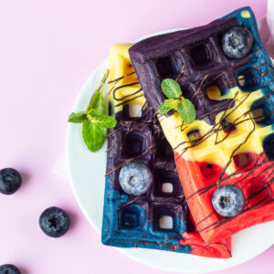 Zwei bunte belgische Waffeln in Rot, Gelb, Blau und Violett auf einem weißen Teller, mit dunklem Schokoladen-Drizzle, frischen Heidelbeeren und Minze
