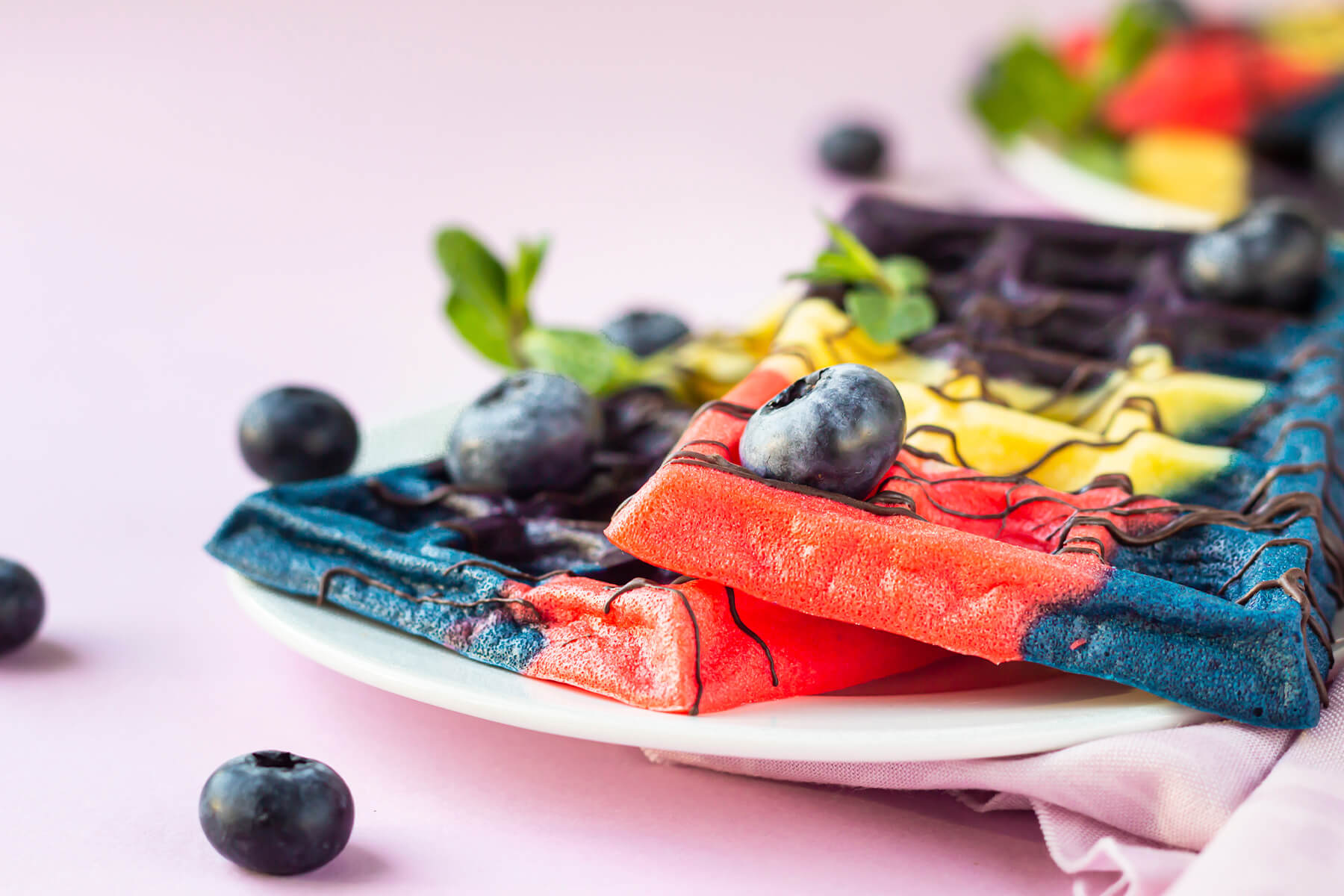 Nahaufnahme einer bunten belgischen Waffel in Blau, Rot und Gelb mit Schokoladenfäden und einer frischen Heidelbeere
