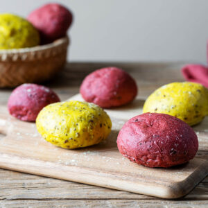 Pinke und gelbe Vollkorn-Brötchen mit Saaten auf einem Holzbrett, im Hintergrund ein Korb mit weiteren bunten Brötchen