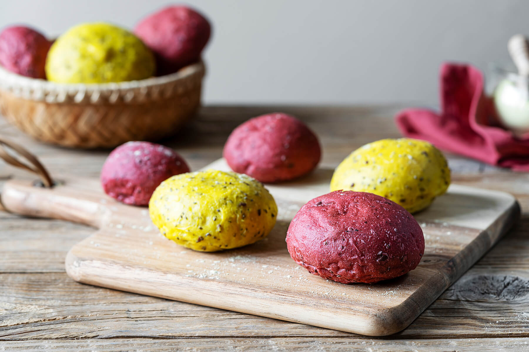 Pinke und gelbe Vollkorn-Brötchen mit Saaten auf einem Holzbrett, im Hintergrund ein Korb mit weiteren bunten Brötchen
