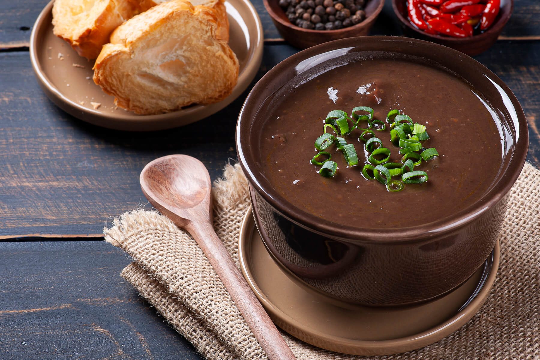 Cremige dunkle Caldo de Feijao aus schwarzen Bohnen in einer braunen Schale mit Frühlingszwiebeln, daneben ein Holzlöffel auf Jutestoff und geröstetes Weißbrot