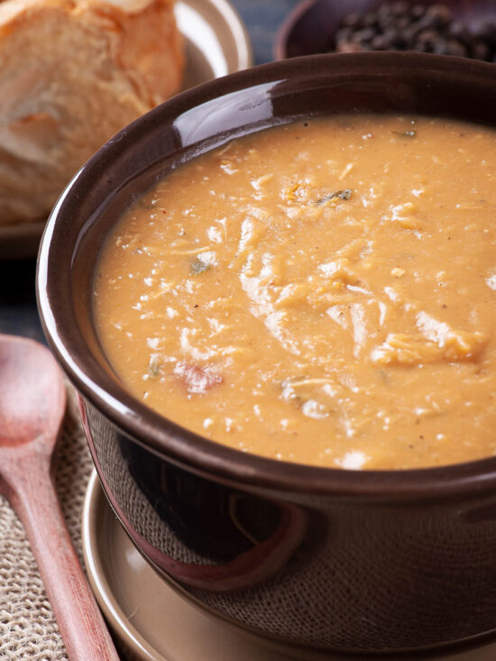 Cremige hellbraune Caldo de Feijao mit zerzupftem Hähnchen in einer dunklen Schale, im Hintergrund Weißbrot und eine kleine Schale mit getrockneten Bohnen