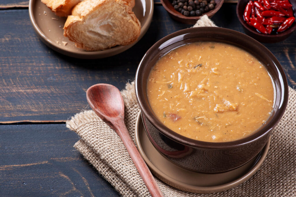 Cremige braune Caldo de Feijao mit zerzupftem Hähnchen und Speck in einer dunklen Schale, dazu Weißbrot, Pfefferkörner und rote Chilis