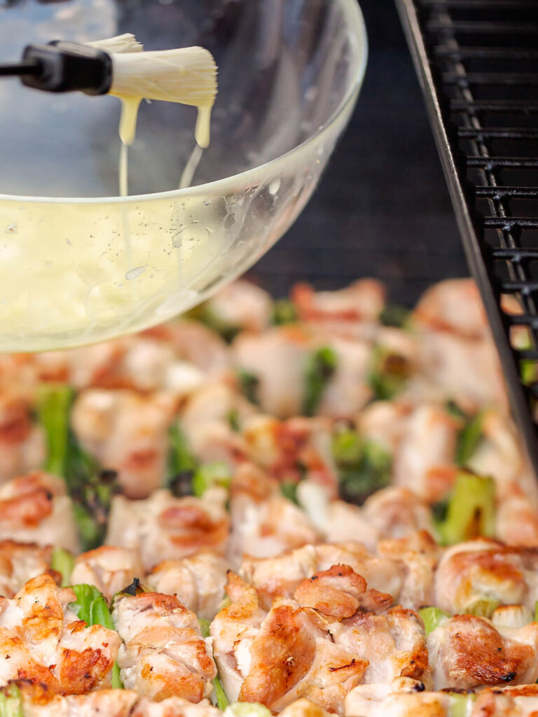 Backpinsel mit heller, zerlassener Butter über einer Glasschüssel, darunter angegrillte Yakitori-Spieße auf dem Grillrost