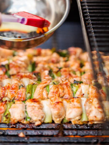 Silikonpinsel und Metallschüssel mit eingekochter Yakitori-Tare über den fast garen Hähnchenspießen auf dem Grill