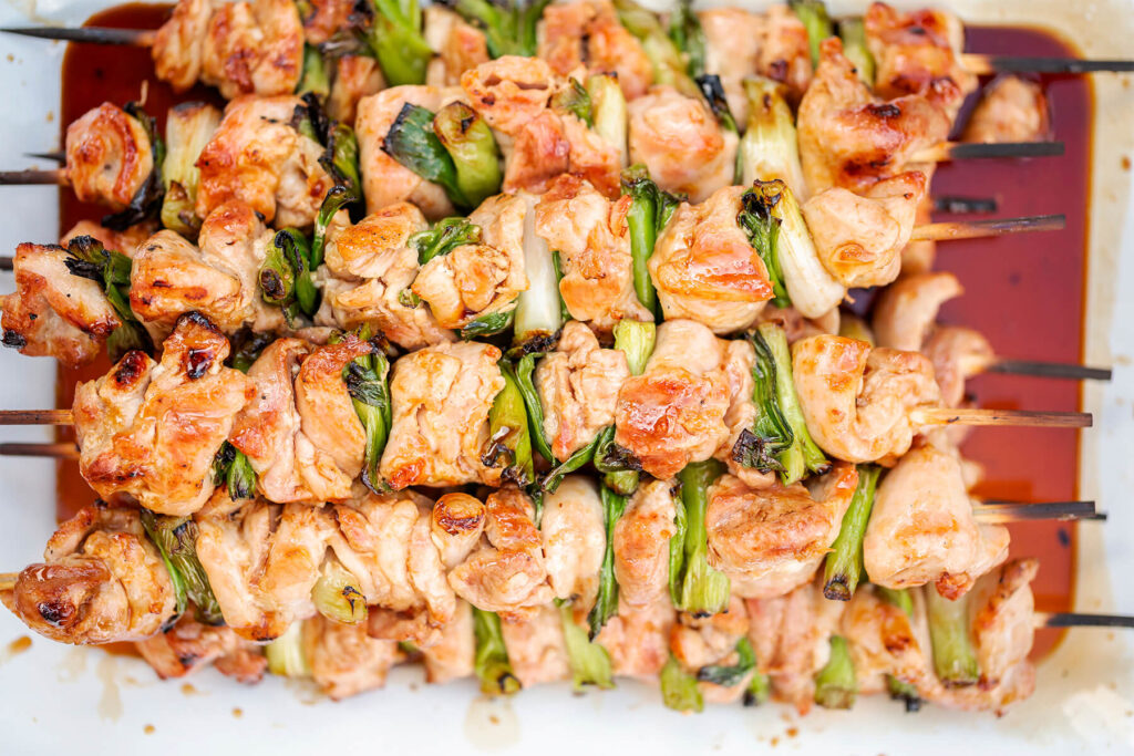 Glasierte Chicken-Yakitori-Spieße mit Frühlingszwiebel von oben auf weißem Blech, hell bernsteinfarben glänzend von der Yakitori-Tare
