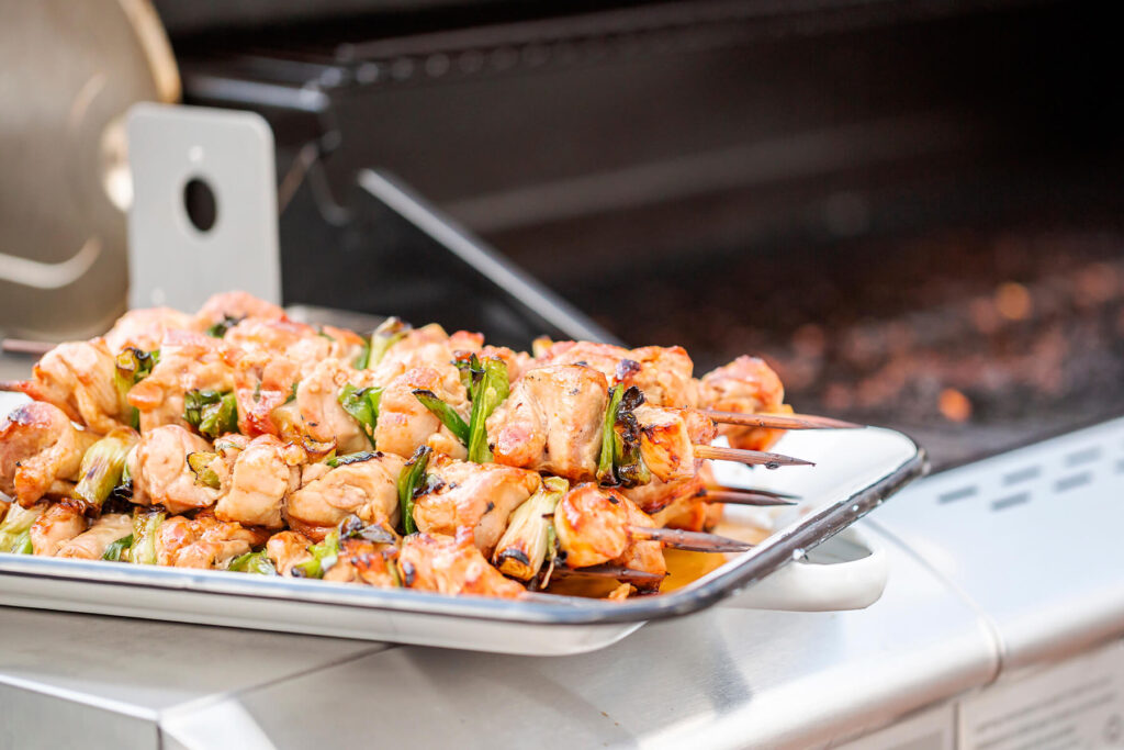 Fertig glasierte Chicken-Yakitori-Spieße auf einem weißen Blech am Rand des Grills, hell bernsteinfarben glänzend von der Yakitori-Tare