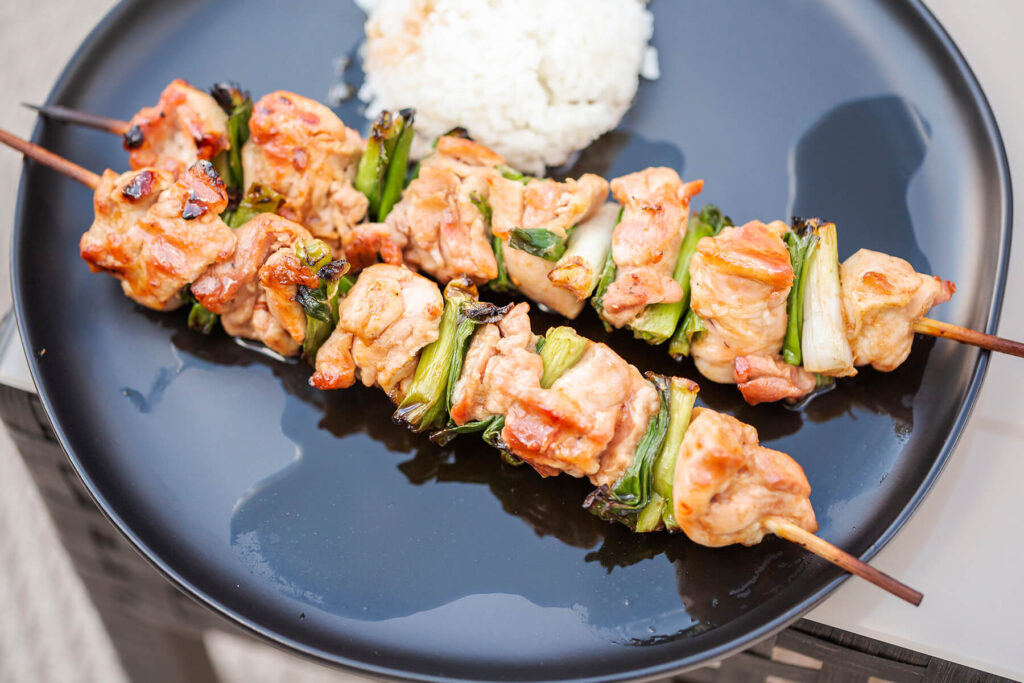 Zwei glasierte Chicken-Yakitori-Spieße mit Hähnchen und Frühlingszwiebel neben einem Häufchen weißem Reis auf einem dunklen Teller