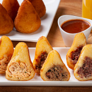 Teller mit drei aufgeschnittenen brasilianischen Coxinhas mit Hähnchen-, Rinderhack- und Carne-Seca-Füllung, dahinter ganze goldbraune Coxinhas, scharfer Dip und Orangensaft
