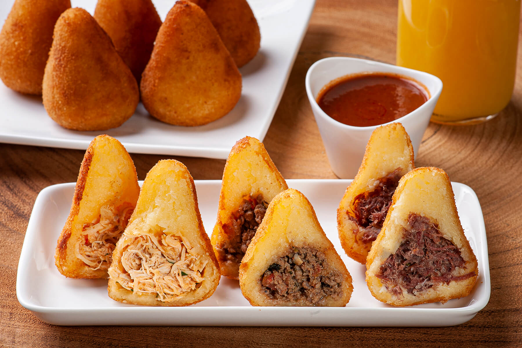 Teller mit drei aufgeschnittenen brasilianischen Coxinhas nebeneinander, gefüllt mit Hähnchen, Rinderhack und Carne Seca, dahinter ganze goldbraune Coxinhas, scharfer Dip und Orangensaft