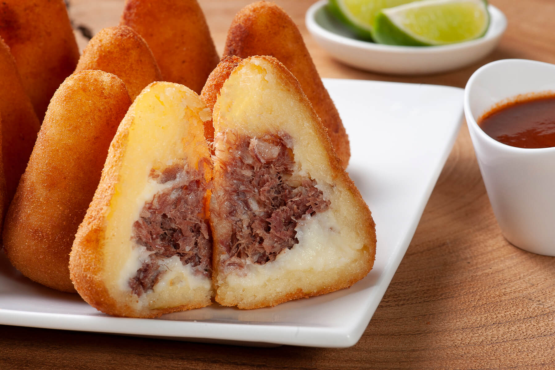 Zwei Hälften einer aufgeschnittenen Coxinha mit Carne-Seca-Füllung auf einem Teller angerichtet, daneben Limettenspalten und scharfer roter Dip