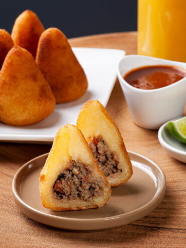 Aufgeschnittene brasilianische Coxinha in Tropfenform mit krümeliger Rinderhackfüllung, goldbraun frittiert, daneben scharfer roter Dip und Orangensaft