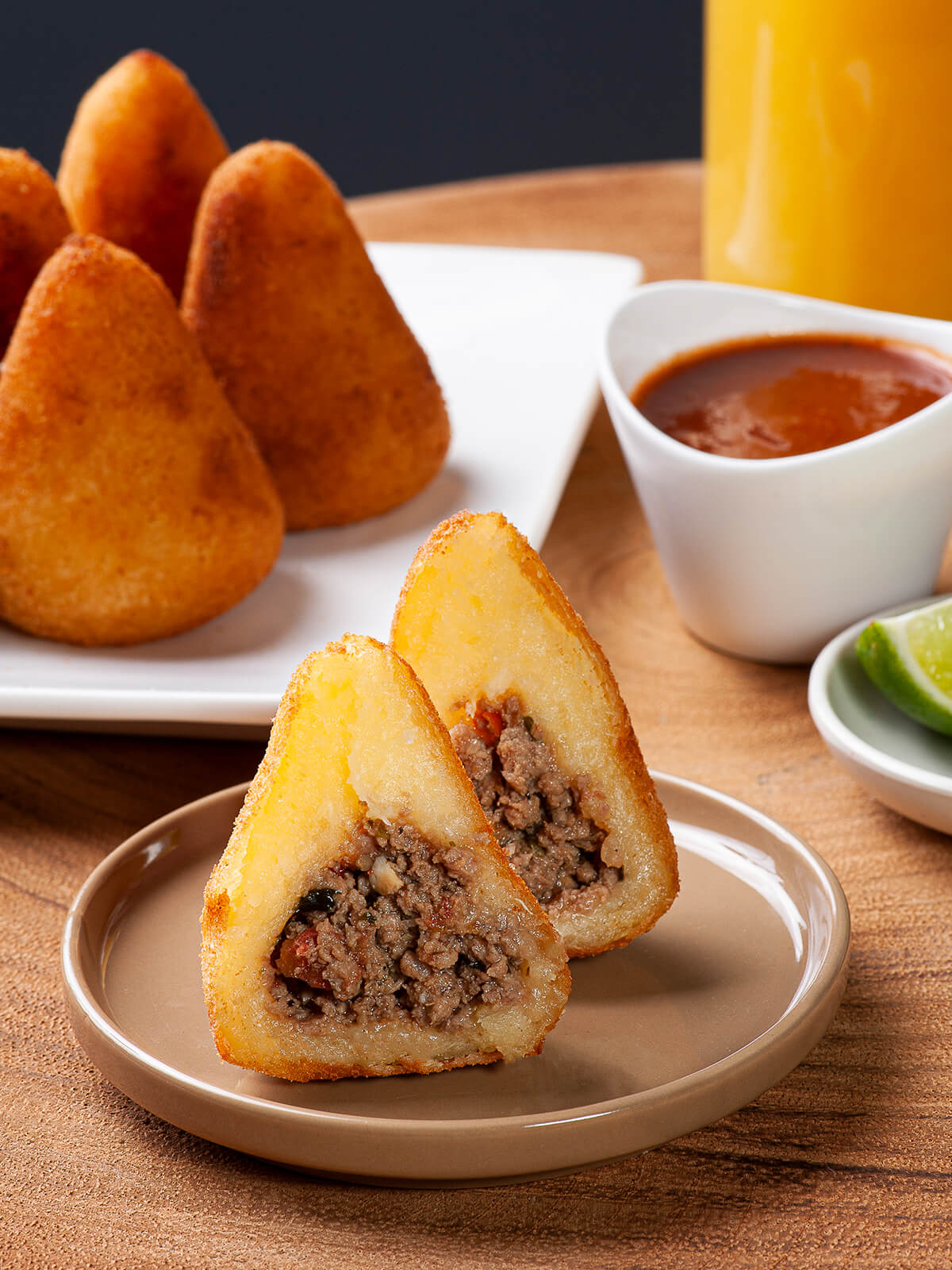 Aufgeschnittene brasilianische Coxinha in Tropfenform mit krümeliger Rinderhackfüllung, goldbraun frittiert, daneben scharfer roter Dip und Orangensaft