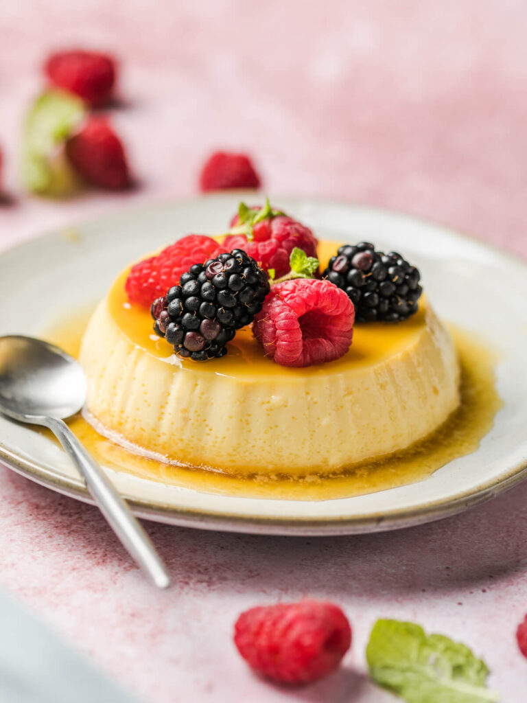 Gestürzte Crème Caramel mit glänzender Karamellschicht auf einem hellen Teller, daneben zwei Löffel und frische Beeren