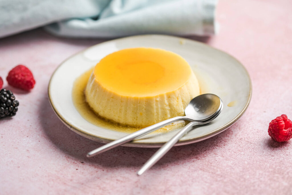 Gestürzte Crème Caramel mit glänzender Karamellschicht auf einem hellen Teller, daneben zwei Löffel und frische Beeren