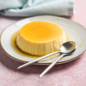 Gestürzte Crème Caramel mit glänzender Karamellschicht auf einem hellen Teller, daneben zwei Löffel und frische Beeren