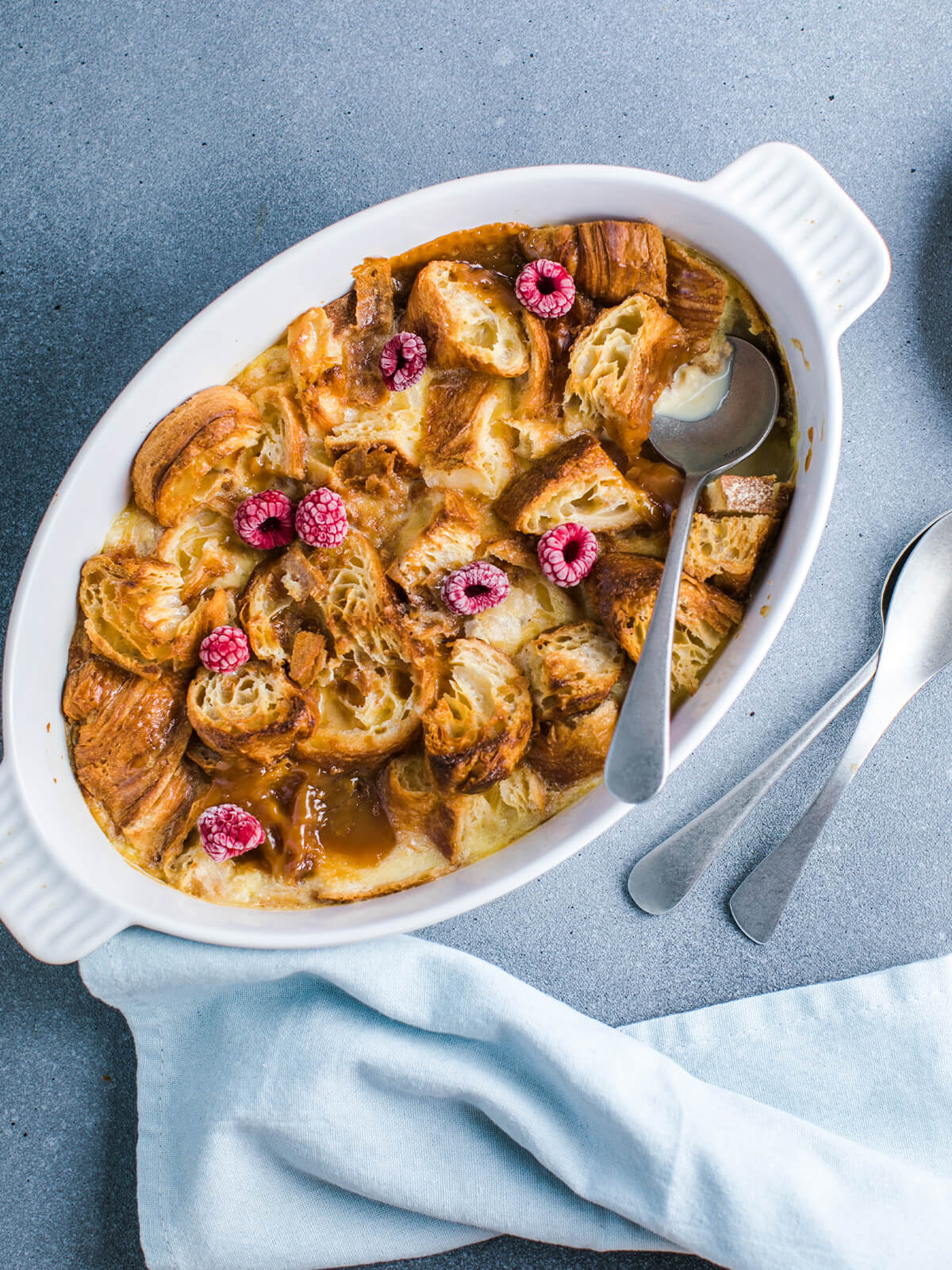 Croissant-Auflauf in weißer ovaler Form mit goldenen Croissant-Stücken, cremiger Vanillesauce und frischen Himbeeren, zwei Löffel daneben