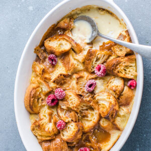 Croissant-Auflauf von oben in weißer ovaler Form, mit cremiger Vanillesauce, goldenen Croissant-Stücken und frischen Himbeeren