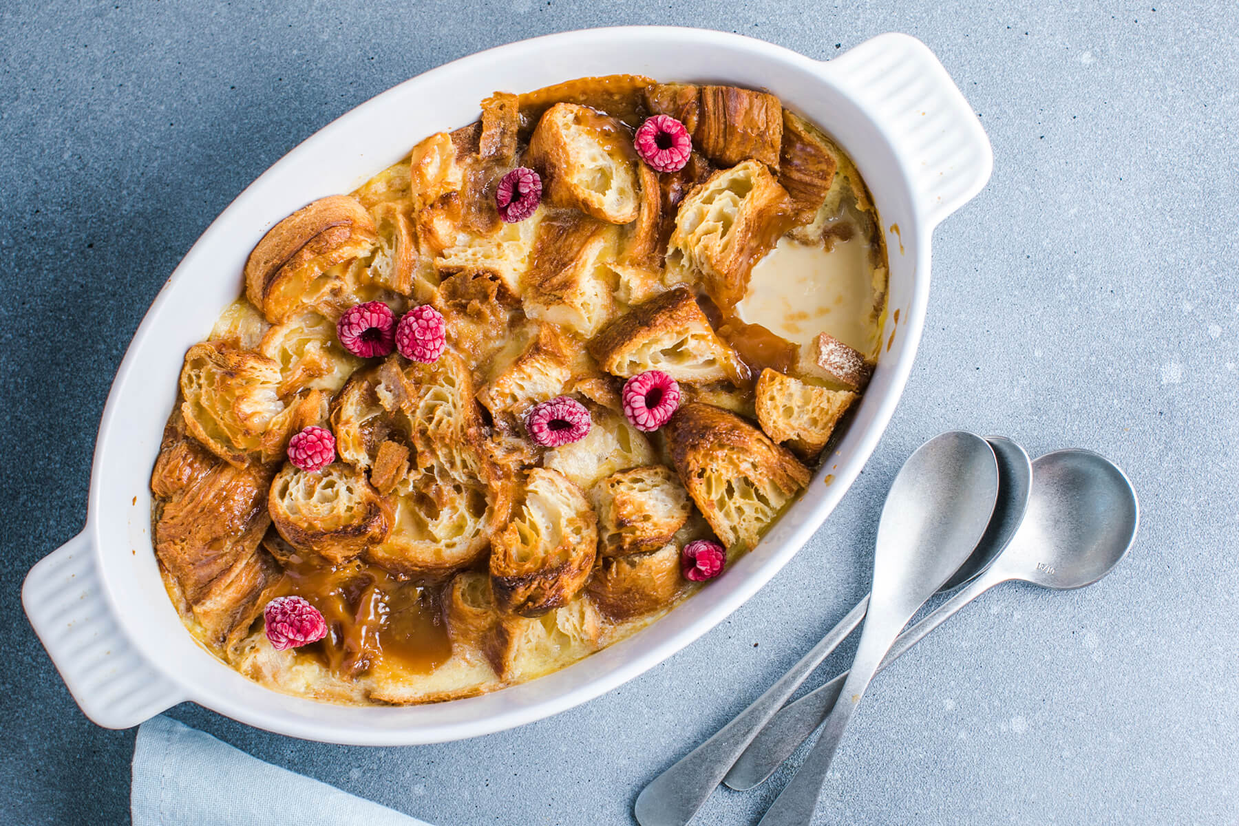 Croissant-Auflauf von oben in weißer ovaler Form mit goldenen Croissant-Stücken, Vanillesauce und Himbeeren