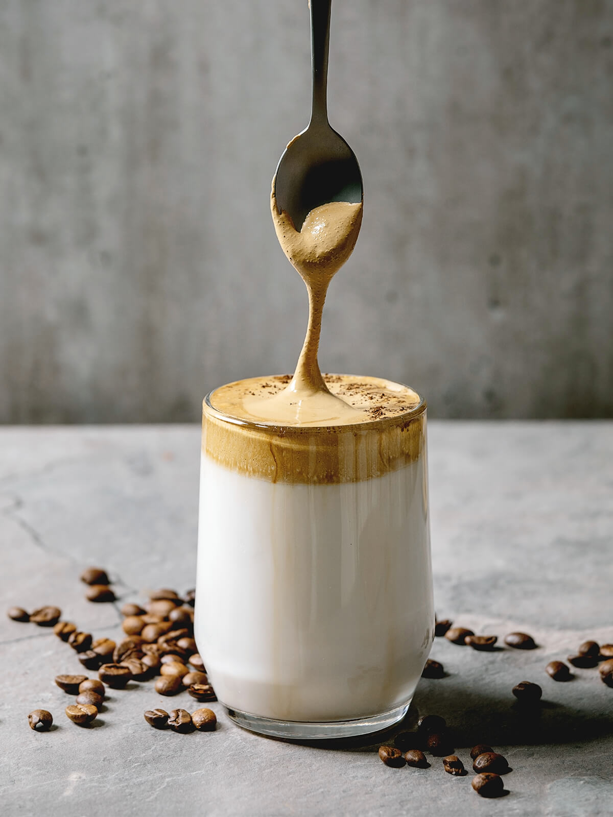 Dalgona-Kaffee im Glas – ein Löffel zieht die dicke, hellbraune Kaffeecreme über die kalte Milch, Kaffeebohnen liegen ringsum
